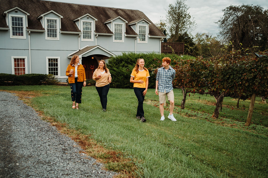 Four people walking and talking through a grassy area near a vineyard, in front of a two-story house with dormer windows. Two women in mustard-colored tops are following a young man in a plaid shirt and a young woman in a yellow blouse.