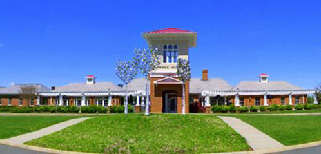 A wide-angle photo shows an expansive building with a tall central tower and multiple roof sections, all with red and grey shingles. It's set against a bright blue sky. The entrance is grand, with a driveway curving around a green lawn in front.