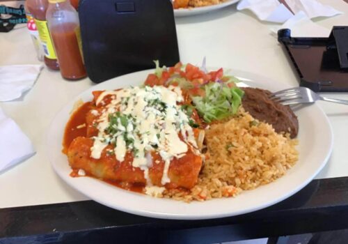 A plate of Mexican food featuring an enchilada topped with red sauce, cheese, and cilantro. The plate also includes a side of refried beans, Mexican rice, and a small salad with lettuce and diced tomatoes. Hot sauce bottles and napkins are in the background. Image
