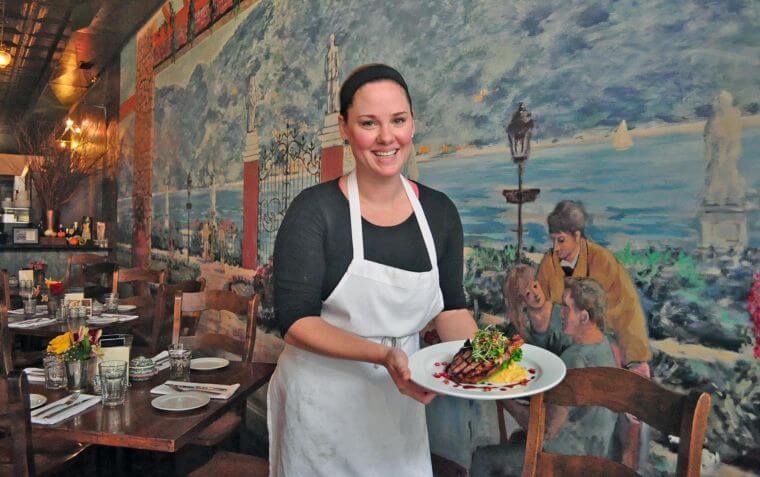 A smiling woman wearing a black shirt, white apron, and headband holds a plate of food. She stands in a warmly lit restaurant with a mural depicting an outdoor coastal scene. Tables are set and decorated with flowers and glasses in the background.