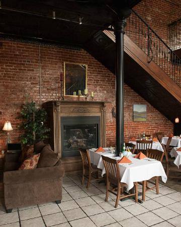 A cozy restaurant with an exposed brick interior, wooden tables covered with white tablecloths, and neatly folded orange napkins. A brown sofa faces a fireplace next to a staircase. Various framed artworks adorn the walls, and a potted plant adds greenery.