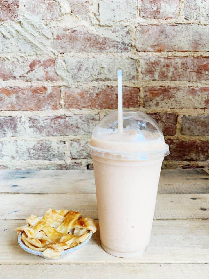 A creamy peach-colored smoothie with a straw in a plastic cup sits on a wooden surface next to a small dish of pie with a flaky crust. The background features a rustic, exposed brick wall.
