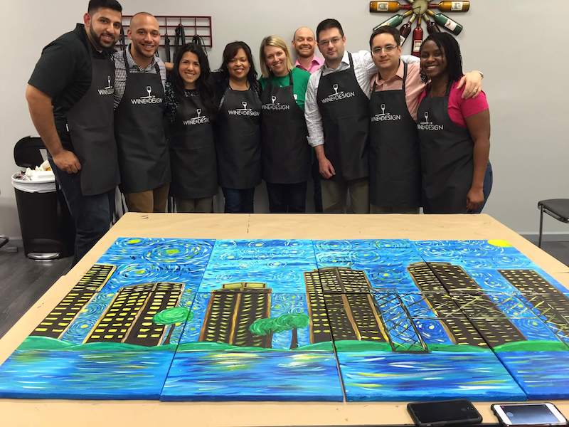 A group of eight people wearing aprons stands behind a table displaying a collaborative painting of a cityscape with swirling blue skies reminiscent of Van Gogh's style. They are smiling and posing for the photo in an indoor setting.