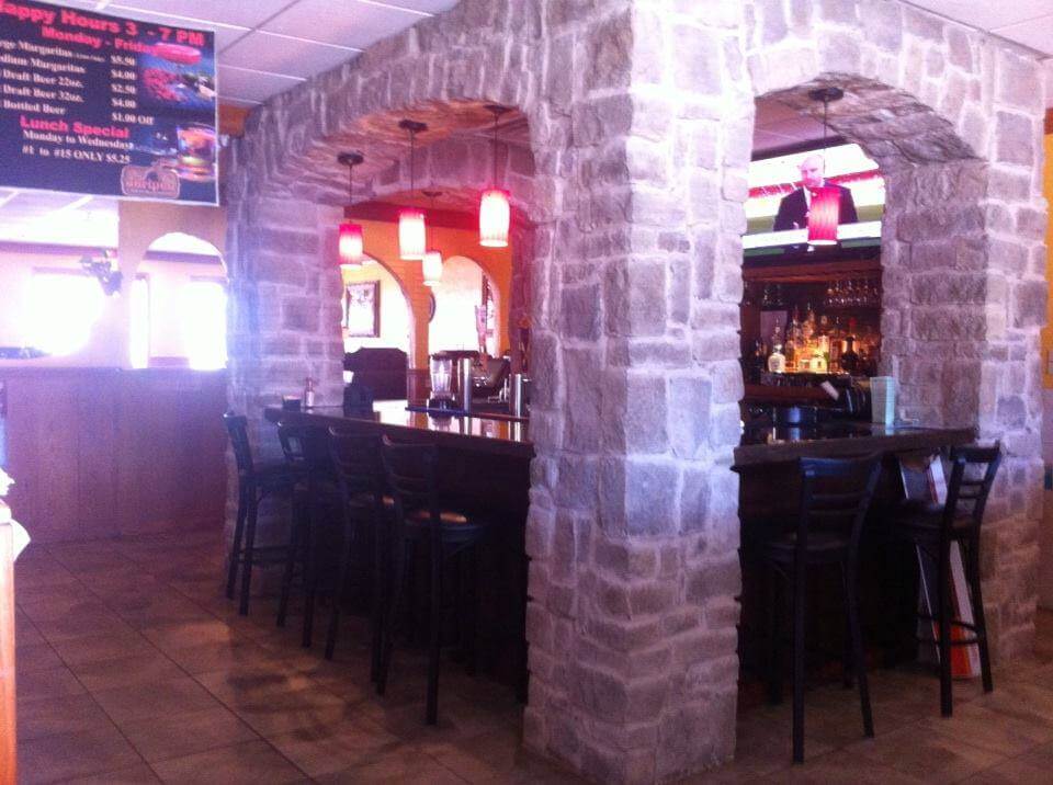 A cozy bar with stone archways and wooden high chairs. The bar is illuminated by red pendant lights hanging from the ceiling. There is a flat-screen TV above the bar, and a menu with happy hour specials is visible on the left side.