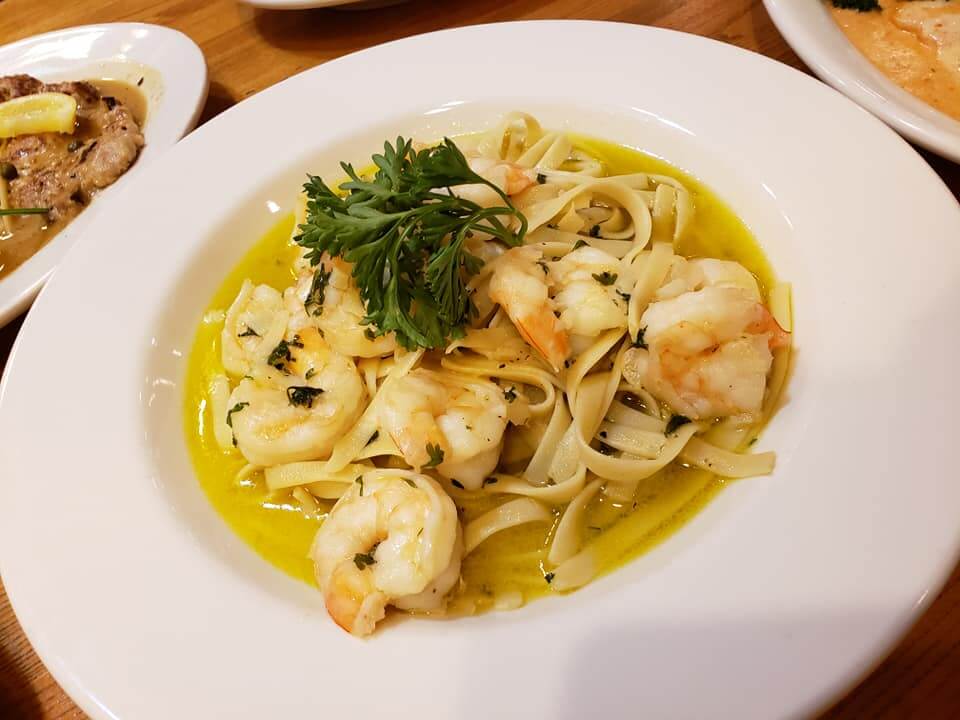A white plate filled with fettuccine pasta, topped with shrimp and garnished with parsley, served in a yellow garlic butter sauce. The dish is placed on a wooden table, with other food items blurred in the background.