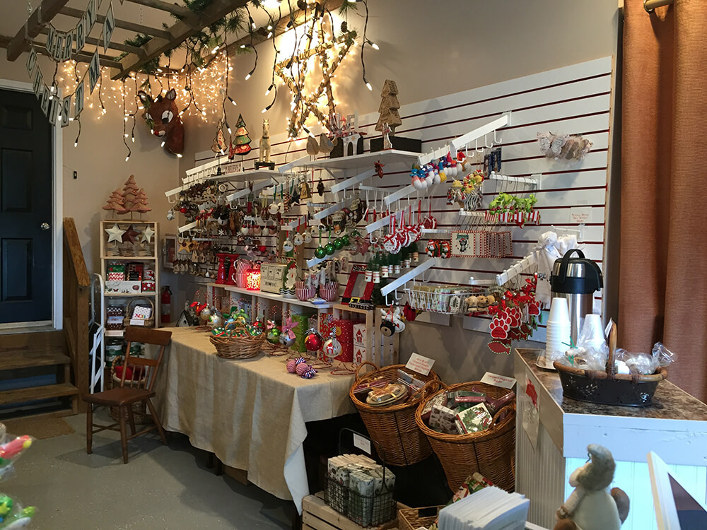 A cozy store brightly decorated with holiday ornaments. String lights and garlands hang from the ceiling, illuminating a variety of colorful decorations on shelves, including ornaments, figurines, and small gift items. A small chair and baskets of goodies are displayed.
