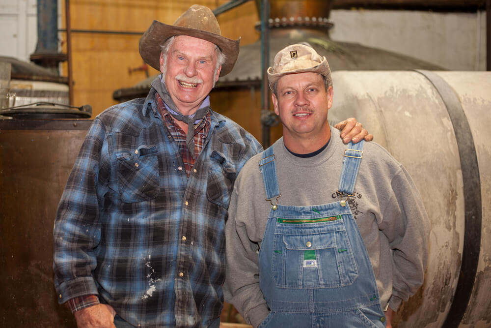 Two men are standing side by side, smiling. The older man on the left wears a cowboy hat, plaid shirt, and bandana. The younger man on the right wears a cap, sweatshirt, and overalls. They appear to be in a workshop or distillery, with large tanks in the background.