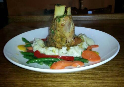 A white plate holds a serving of mashed potatoes topped with a thick cut of bone-in meat, likely a lamb shank, garnished with herbs. Surrounding the dish are colorful steamed vegetables, including green beans, carrots, and bell peppers. The plate rests on a wooden table. Image