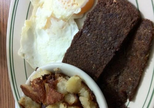 A plate with two fried sunny-side-up eggs, two slices of dark brown bread or possibly meat, and a bowl of fried potato chunks. The food is set on a newspaper, with part of a cup visible in the bottom left corner. Image
