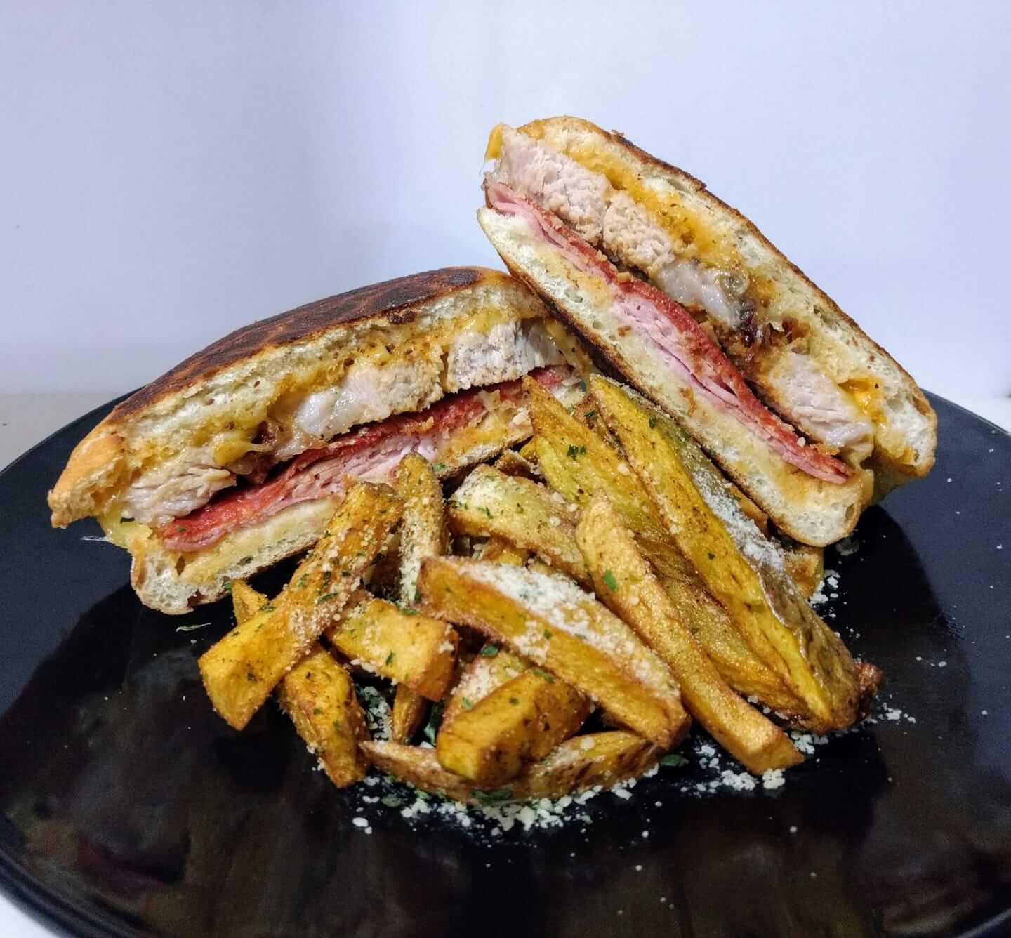 A grilled sandwich cut in half, filled with melted cheese, sliced meat, and crispy chicken, is placed on a black plate. It is served with a side of seasoned golden-brown potato fries garnished with herbs and grated cheese.