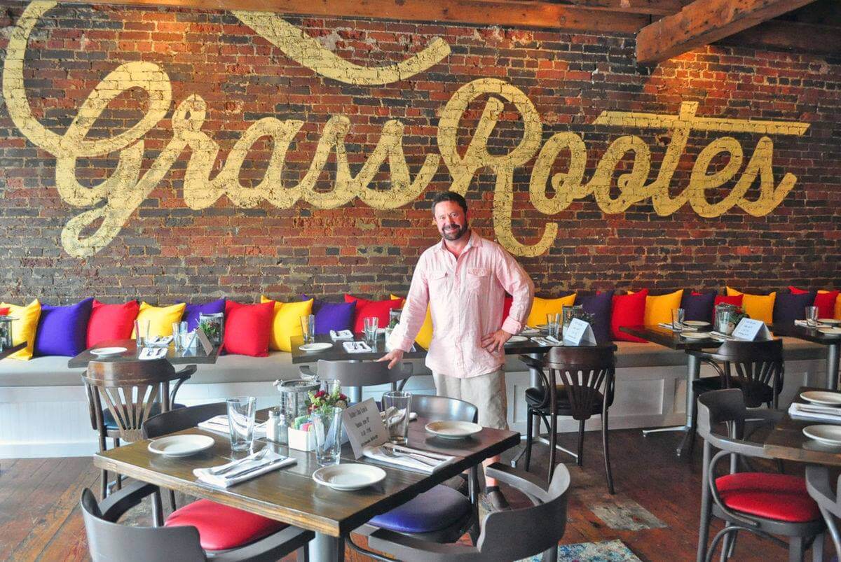 A man in a light pink shirt stands smiling in the dining area of a restaurant called 