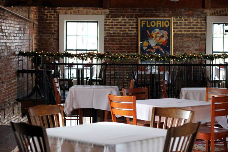 A cozy restaurant interior with brick walls and wooden furniture. Tables are covered with white tablecloths, and a garland with lights hangs along a railing. A 
