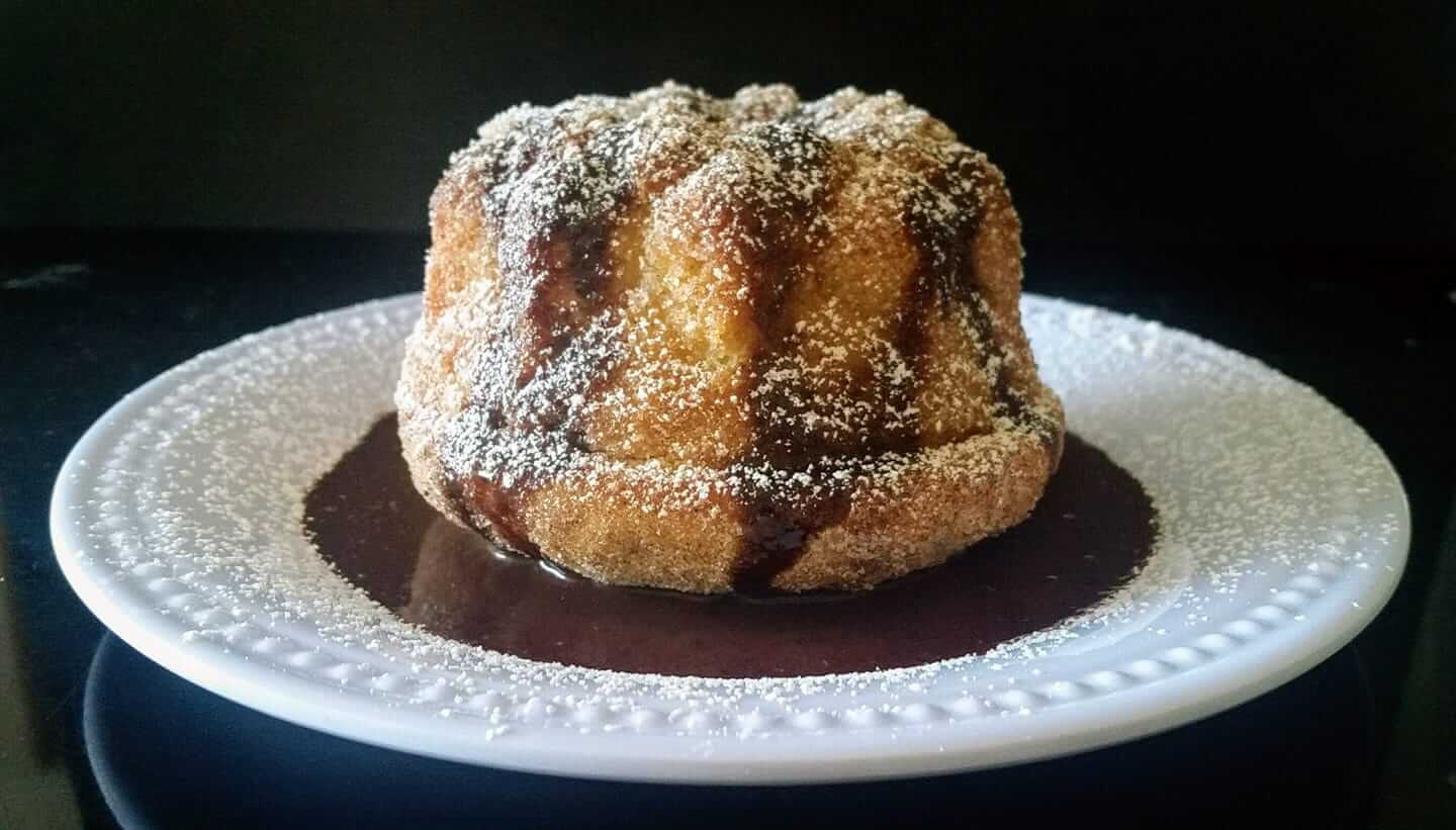 A sugared bundt cake with drizzled chocolate sauce sits on a white plate dusted with powdered sugar. The dark background contrasts with the golden brown cake, highlighting its texture and rich appearance.