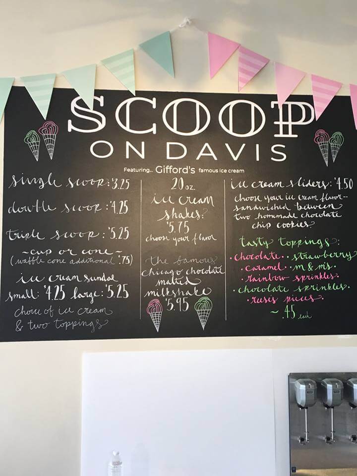 A chalkboard menu at Scoop on Davis ice cream shop. It lists prices for single, double, and triple scoops, ice cream sundaes, 20 oz. ice cream shakes, hot chocolate, and toppings. Colorful pastel bunting decorates the top of the board.