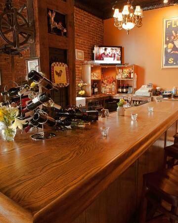 A cozy bar with a polished wooden counter, four shot glasses, and decorative wine bottles. A TV is mounted on a brick wall, and shelves behind the counter hold various bottles and glasses. A chandelier hangs from the ceiling, and a framed butterfly picture decorates a column.