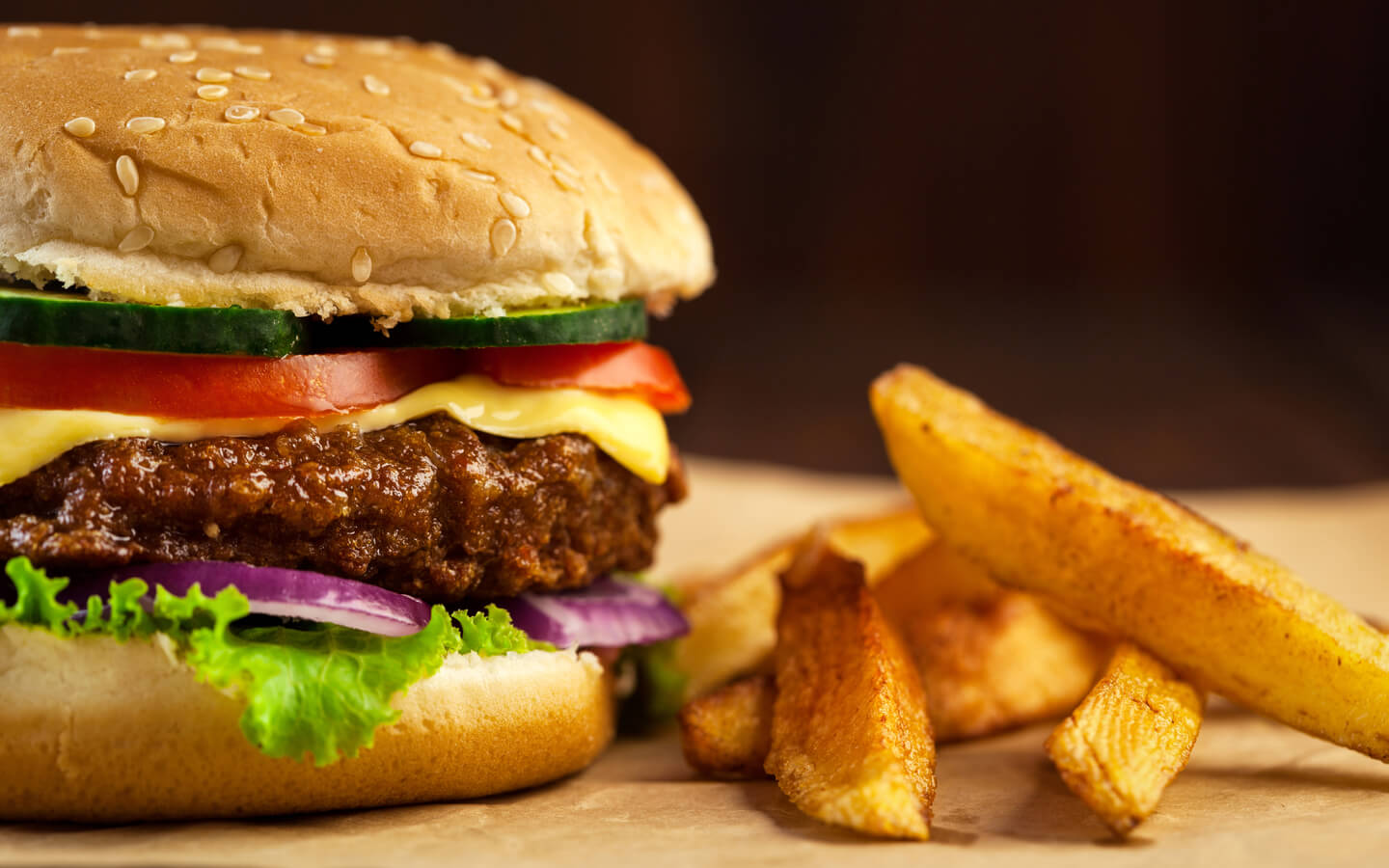 A close-up view of a gourmet cheeseburger with a sesame seed bun, topped with lettuce, red onions, a beef patty, melted cheese, tomato, and cucumber slices. To the right, a serving of thick-cut, golden-brown fries rests on a piece of brown parchment paper.