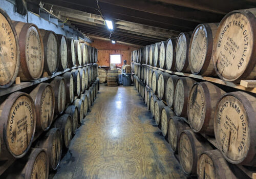 A narrow hallway in a distillery lined with large, wooden barrels stacked on either side. The barrels are marked with 