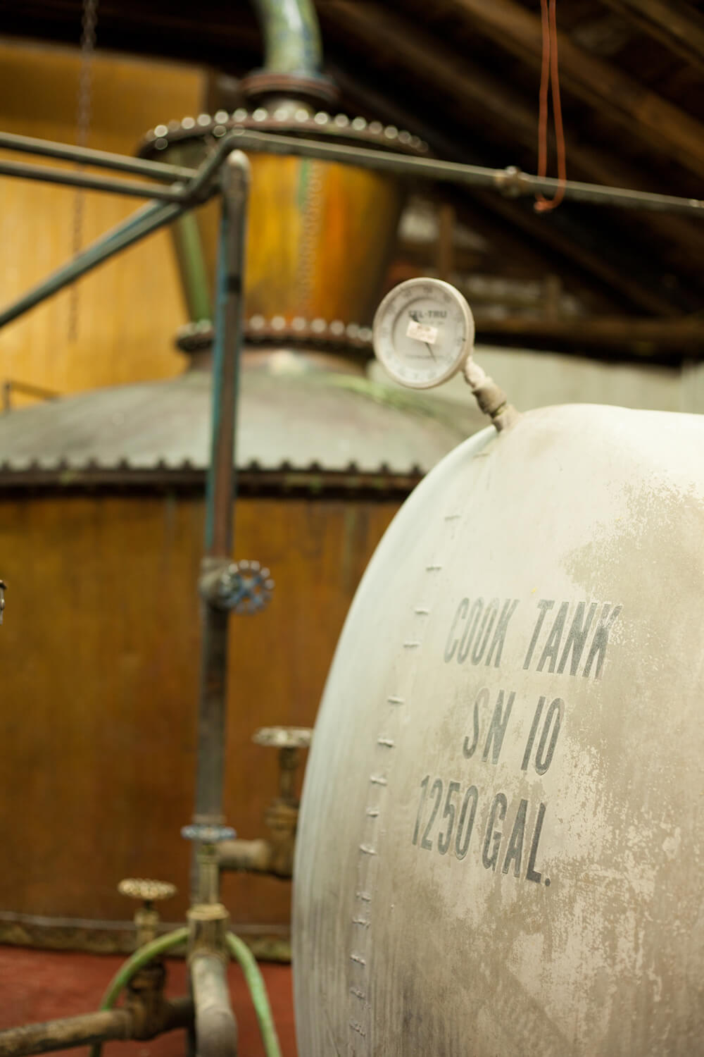 A close-up of industrial equipment in a distillery. The foreground features a large metal tank labeled 
