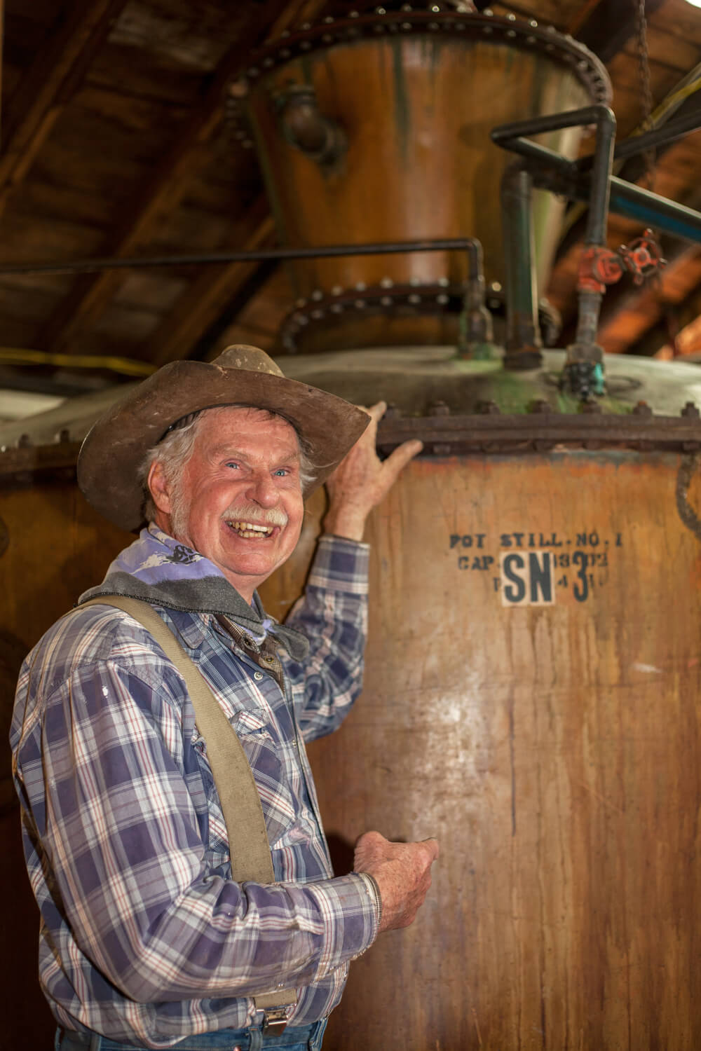 An older man with a broad smile, wearing a cowboy hat, plaid shirt, and suspenders, stands in front of a large, rustic distillery pot still labeled 