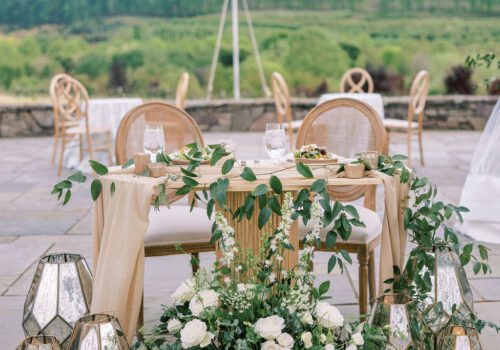 An elegant outdoor wedding table is adorned with a cream tablecloth, lush greenery, and white floral arrangements. Surrounded by decorative lanterns, the table faces an open field with string lights above and several wooden chairs in the background. Image