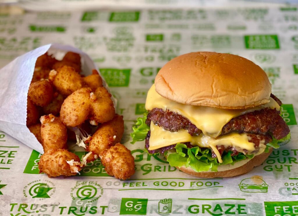 A cheeseburger with lettuce and double patties sits on a wrapper with green text. Next to it is a side of fried cheese curds in a white paper bag. The background wrapper features various green phrases and logos related to the food brand.