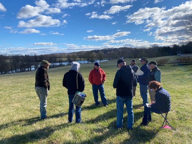 A group of people wearing jackets and hats stand and talk outdoors. It's a sunny day with scattered clouds. Behind them are fields, some trees, and a body of water. One person sits on a small stool, taking notes.
