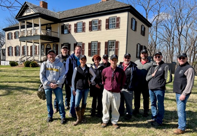 A group of eleven people, both men and women, stand outside in front of a large historic house with beige siding and dark shutters. The group is dressed in casual outdoor attire and appears to be smiling for the photo on a sunny day.