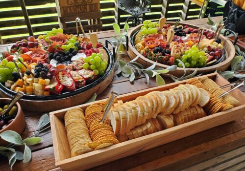 A rustic wooden table displays an assortment of foods: a rectangular tray with various crackers and sliced baguettes, and two circular platters filled with grapes, berries, cheese cubes, stuffed peppers, and charcuterie, garnished with greens and wooden picks. Image