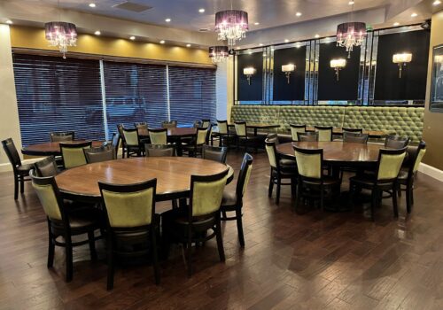 A dining room with several round wooden tables and cushioned chairs arranged neatly. The room features modern chandeliers, wall sconces, and dark wooden flooring. Large windows with blinds are on one side, and a tufted green banquette lines another wall. Image