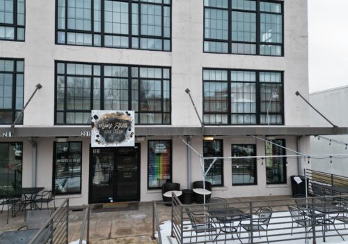 A street view of a modern building with large grid windows. The entrance has a sign reading 