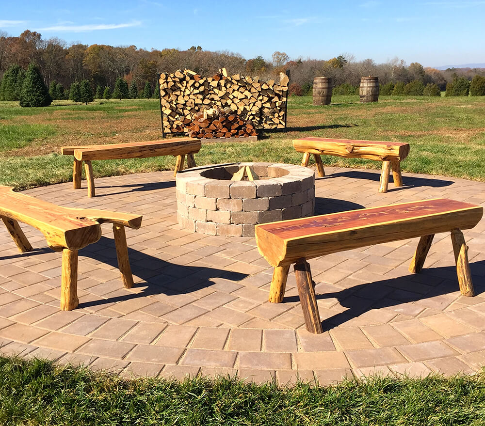 A circular brick fire pit is surrounded by four rustic wooden benches on a paved area. In the background, stacks of firewood are neatly arranged against a metal grid with fields, trees, and a clear blue sky.