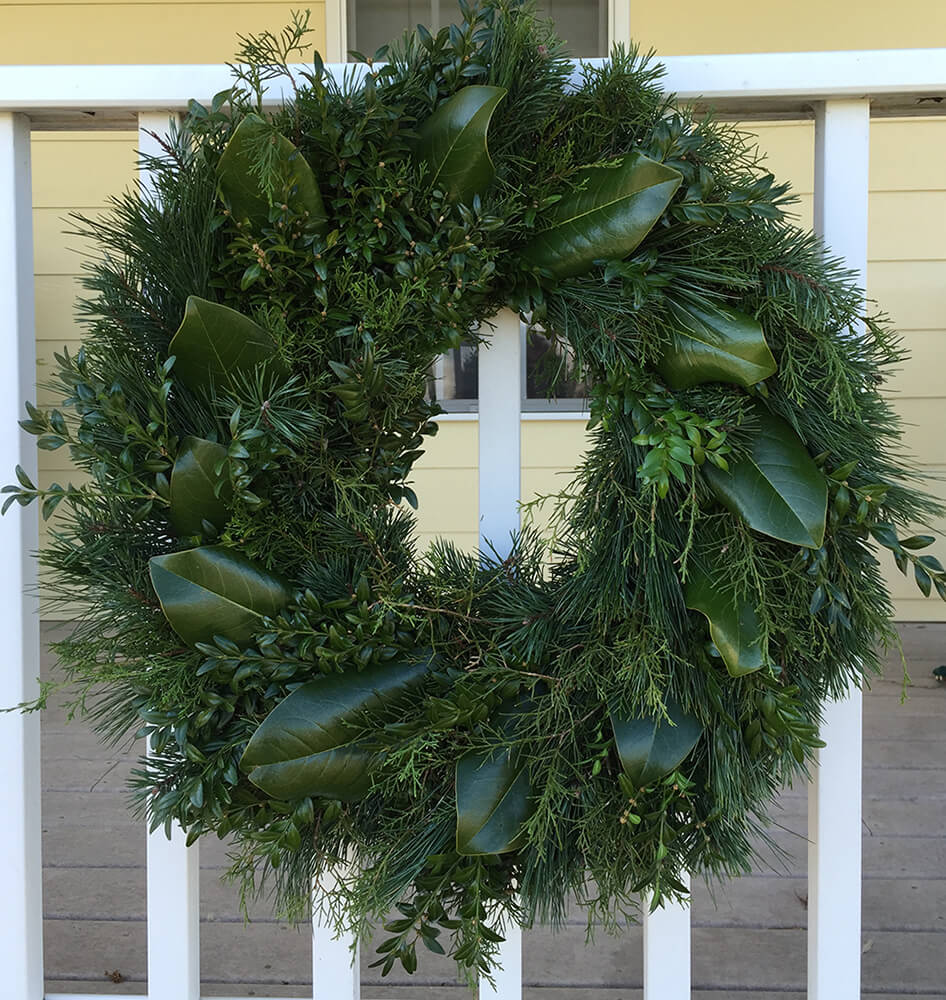 A lush green wreath adorned with various types of foliage, including thick leaves and delicate pine needles, hangs on a white railing in front of a beige-colored house.