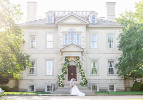 A bride and groom embrace in front of a large, elegant mansion adorned with flower garlands around the entrance. The mansion is off-white with large windows and symmetrical architectural features. Sunlight filters through the trees, adding a soft glow to the scene. Image