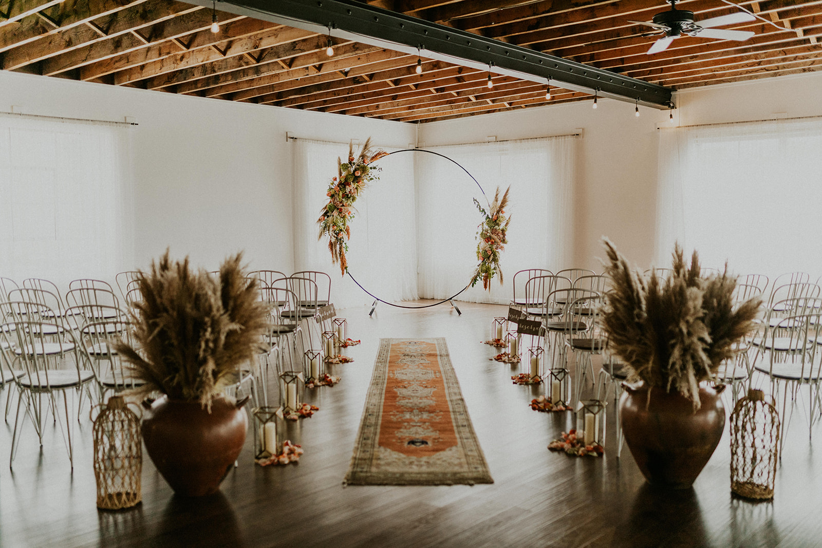 A minimalist indoor wedding setup at our premier wedding and event venue in Culpeper Virginia features a circular floral arch at the front, white walls, and a wooden ceiling. Rows of clear chairs line the aisle, adorned with a patterned rug. Elegant pampas grass in large vases and lanterns complete the look.