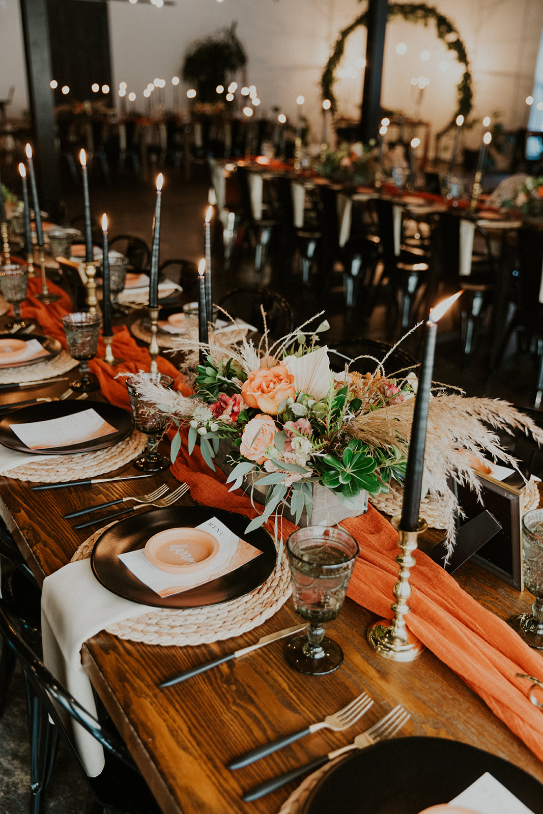 A dining table set for a formal event at our wedding and event venue in Culpeper, Virginia features black plates, woven placemats, and orange napkins. Black candles in brass holders are placed along the center with a floral arrangement of pink roses and greenery. Soft ambient lighting creates a warm, elegant atmosphere.
