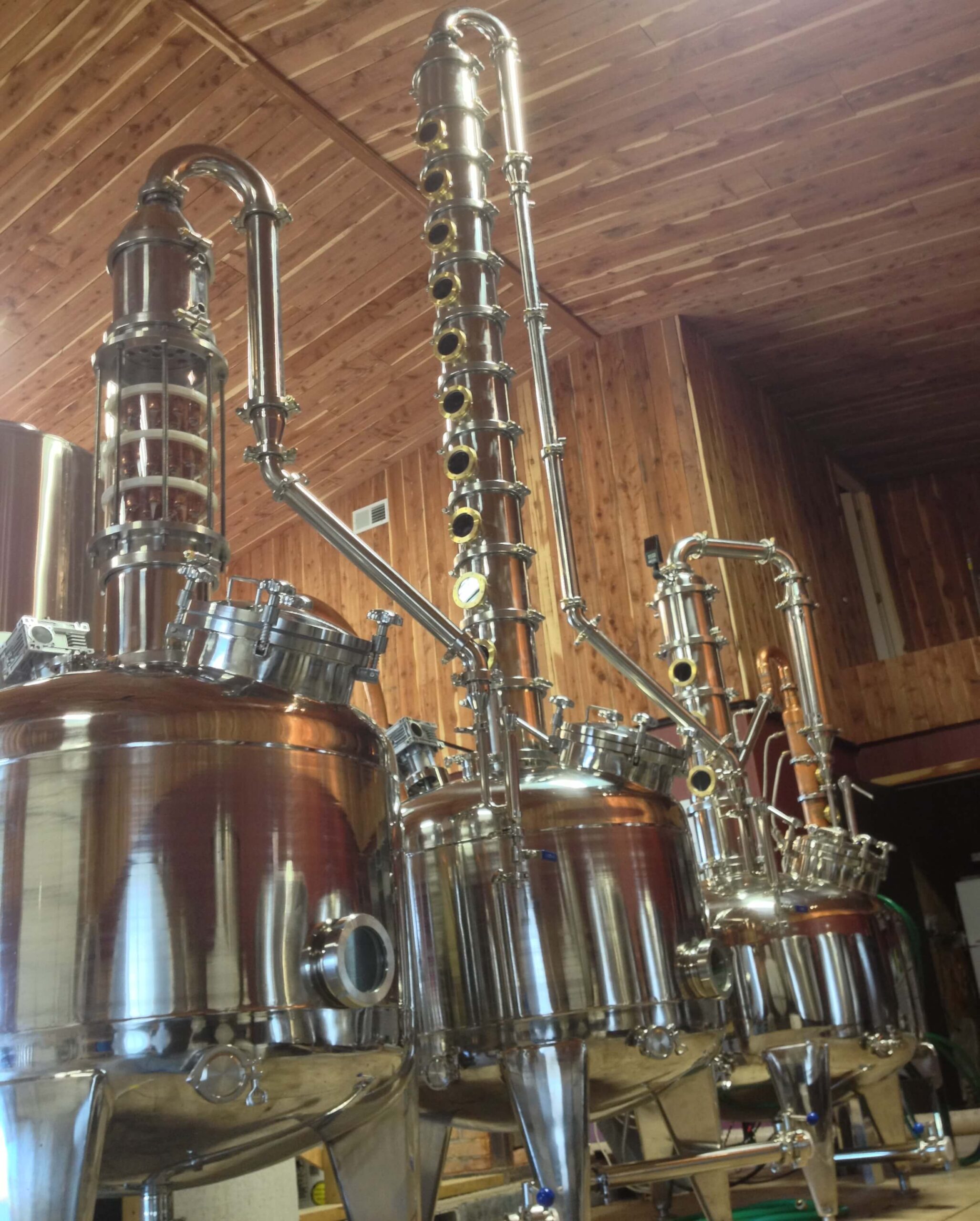 A distillery interior featuring several polished stainless steel distillation columns and tanks, with intricate piping and valves. The wooden walls and ceiling provide a rustic backdrop to the industrial equipment.