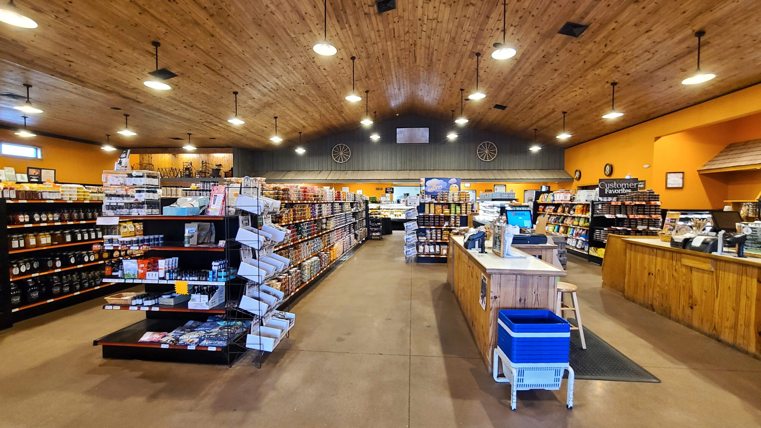 A spacious store interior with wooden ceilings and brightly lit shelves filled with a variety of products, including groceries and household items. The store features a central checkout counter and organized aisles, reflecting a rustic, inviting atmosphere.