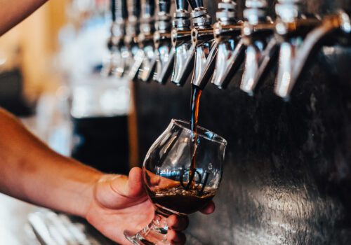 A person is filling a glass with dark beer from a tap at a bar. The beer tap system features multiple dispensers lined up in a row. The glass is held at an angle to minimize foam. The scene suggests a lively bar setting. Image