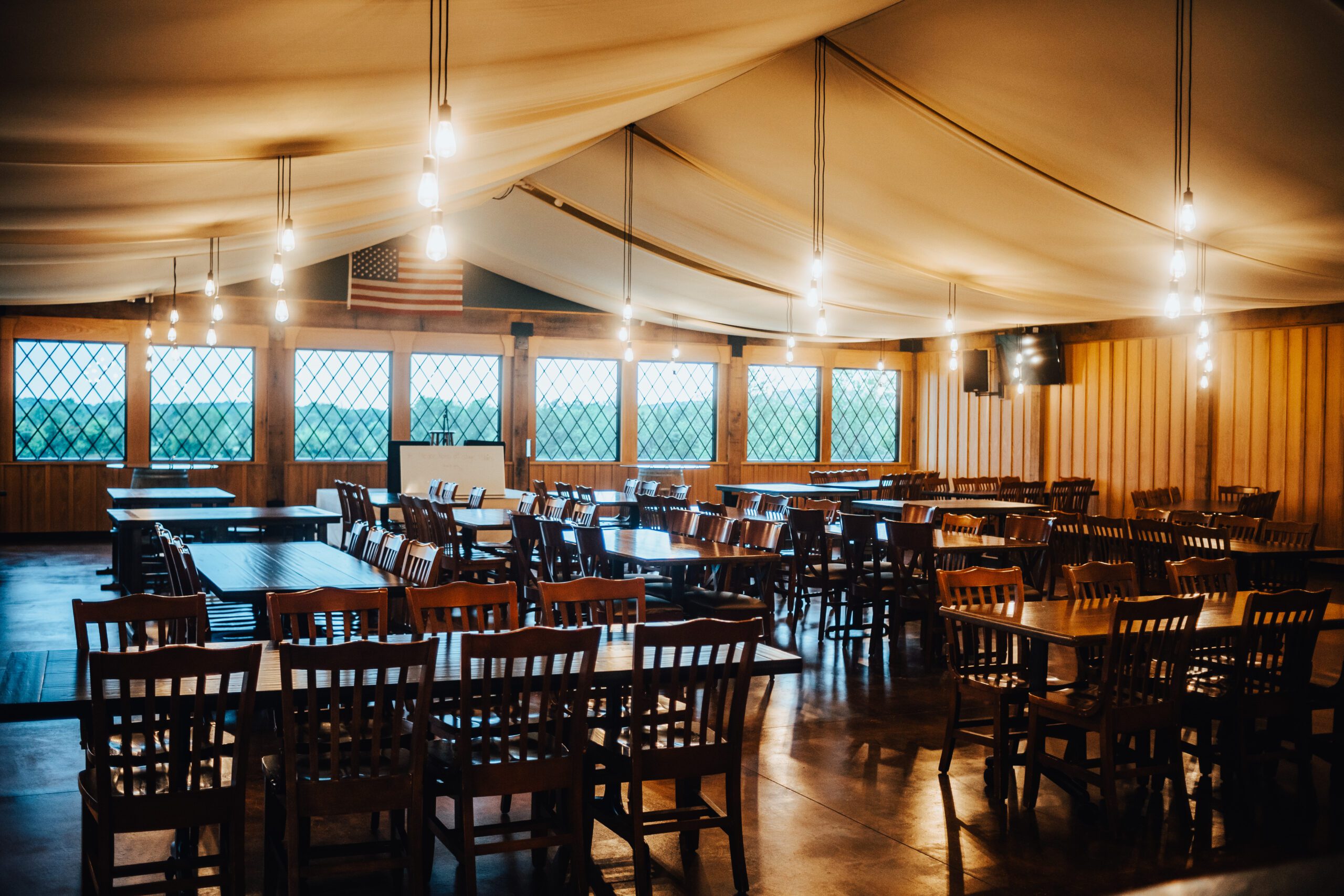 A spacious dining hall with wooden tables and chairs arranged neatly. The ceiling is draped with white fabric and hanging lights. Diamond-paned windows line the far wall with a view of greenery outside. An American flag hangs on one wall.
