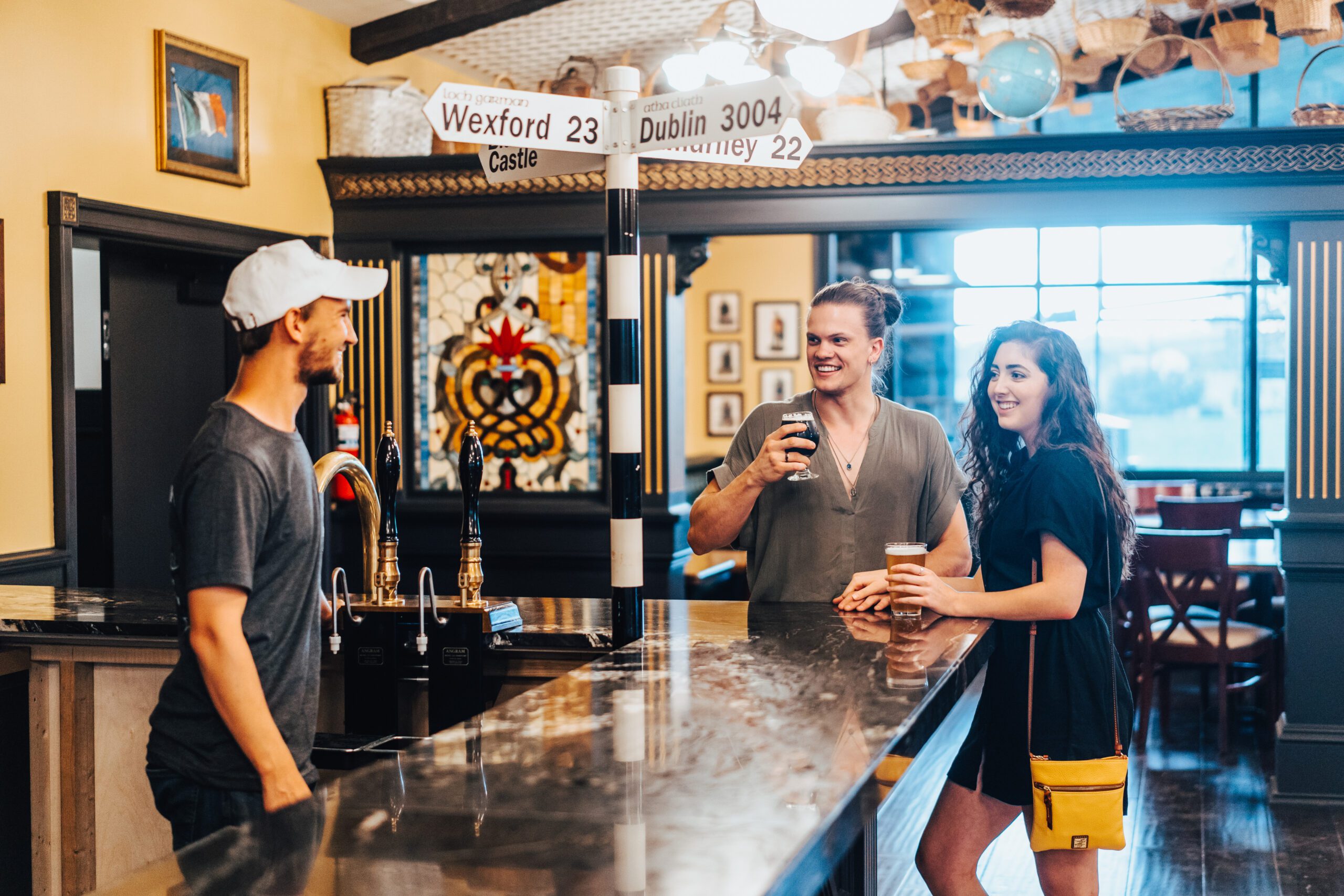 Three people are at a bar; a man behind the counter, and a man and woman standing in front of it, holding drinks. They are smiling and engaged in conversation. Behind them is a stained-glass window and a signpost with directional signs and distances to Wexford, Dublin, and other places.