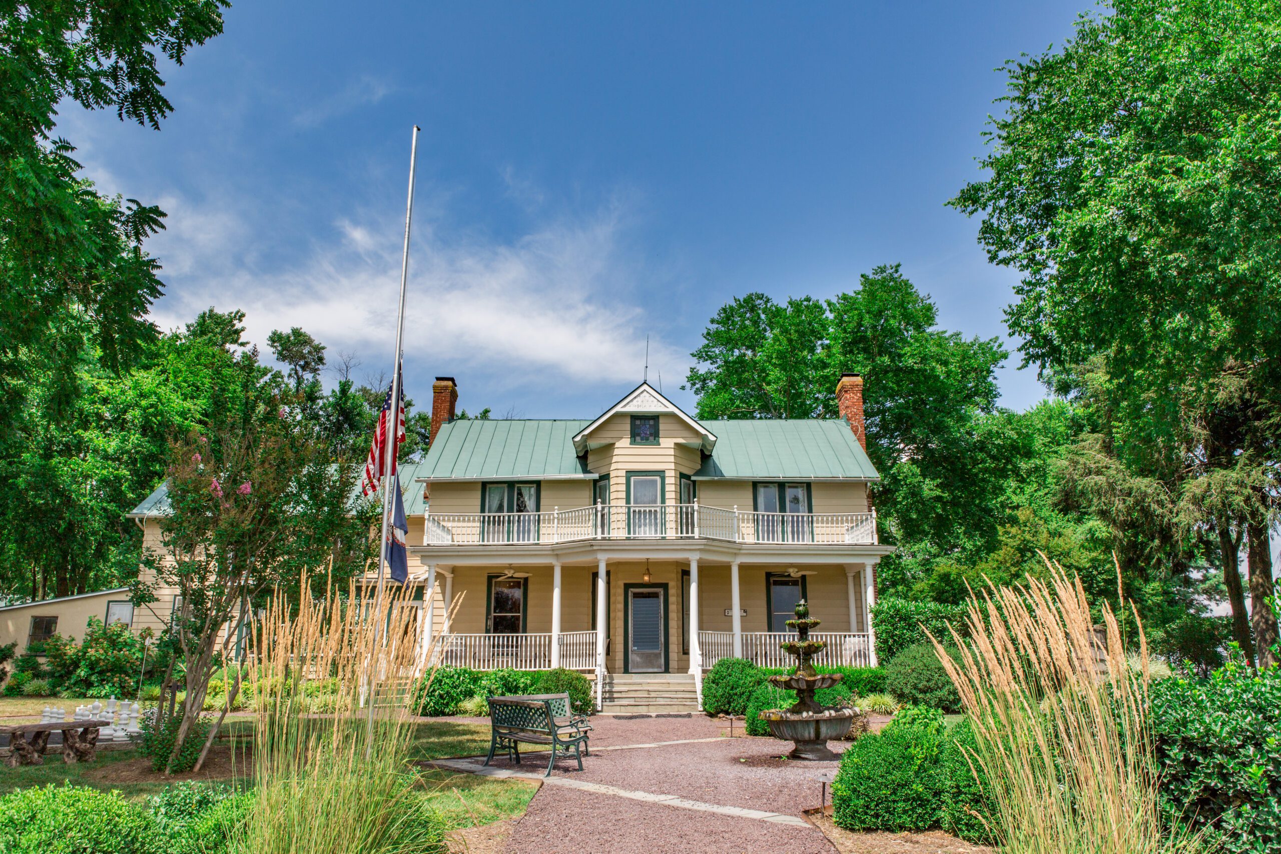 A large, two-story house with beige siding and a green roof stands amidst lush greenery. It features a central front door with a covered porch and a balcony above. An American flag at half-mast is in front, and there's a pathway leading to the entrance, flanked by benches and manicured bushes.