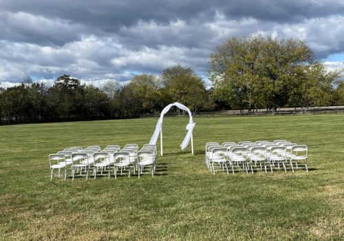 An outdoor wedding setup features rows of white chairs arranged on a green lawn. A simple wooden arch draped with white fabric stands at the front, set against a backdrop of trees and a partly cloudy sky. Image