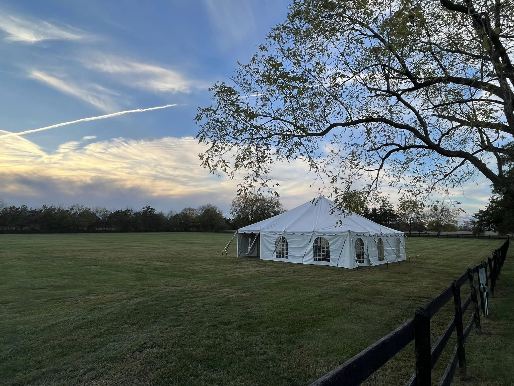 A white event tent is set up in a large, open grassy field near a wooden fence. A tree with sparse leaves stands nearby. The sky is partly cloudy with streaks of sunlight and contrails, creating a serene, picturesque scene.