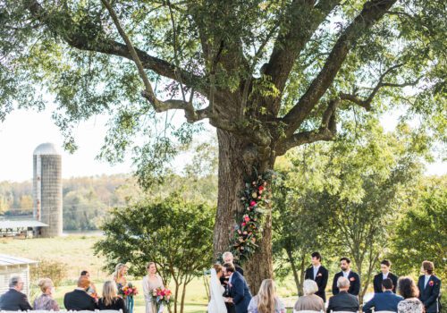 A wedding ceremony takes place outdoors under a large tree adorned with flowers. The bride and groom stand in front of an officiant, surrounded by the wedding party. Guests are seated on white chairs, with a rustic farm scene and silo in the background. Image