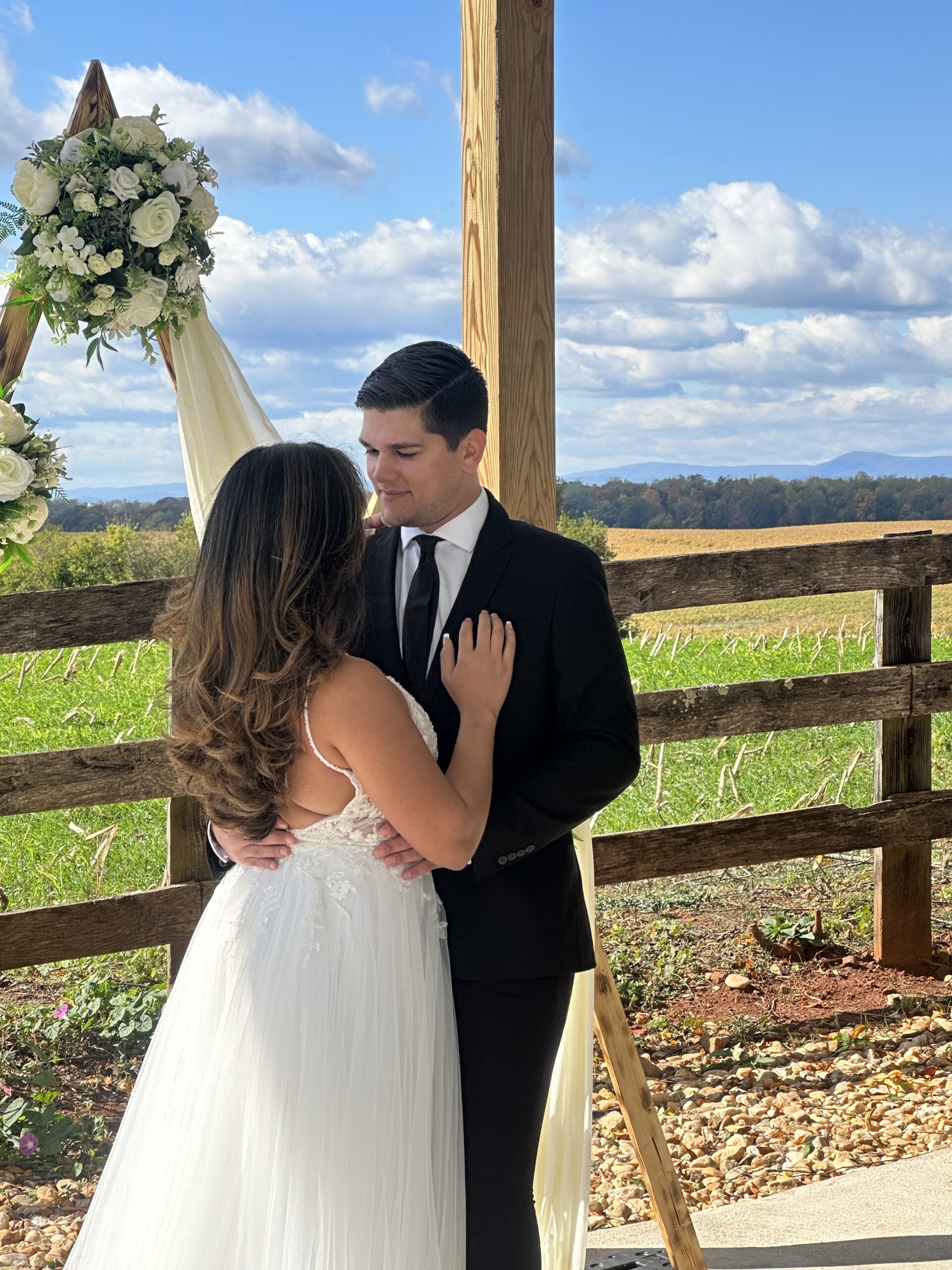 A bride and groom stand close together under a wooden arch adorned with white flowers and draped fabric. They gaze into each other's eyes, with the green fields and blue sky with clouds in the background. The bride wears a white dress, and the groom is in a black suit.