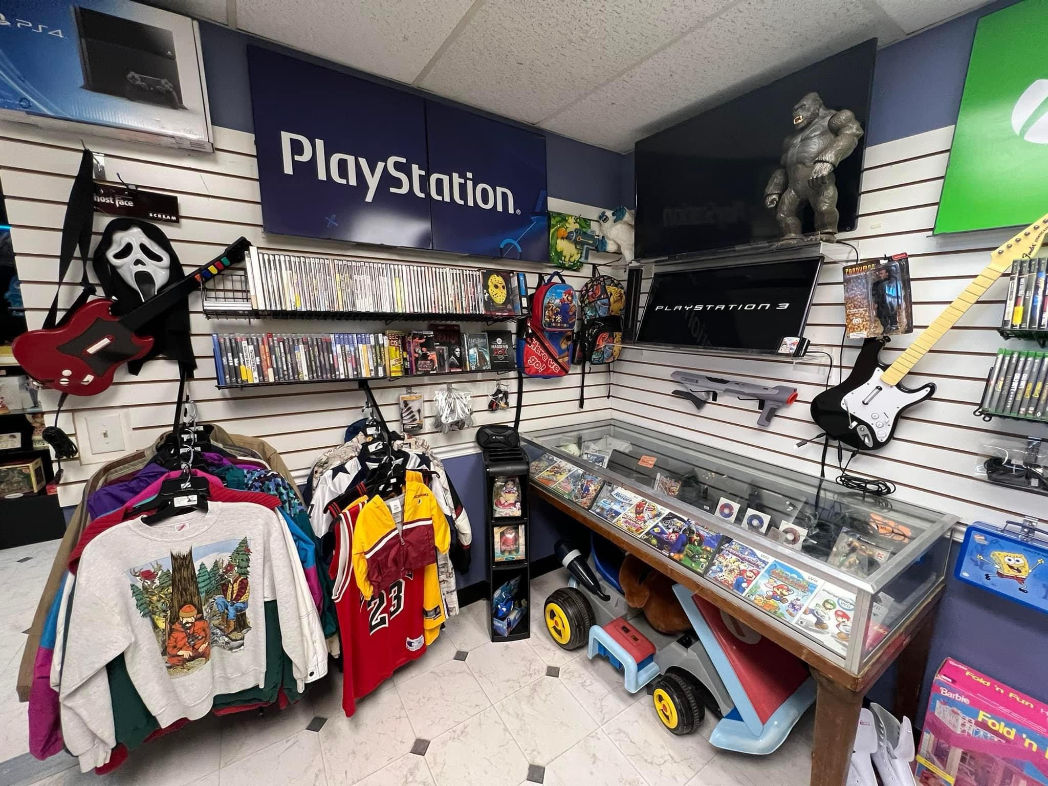 A retro gaming store displays an array of PlayStation games on shelves, alongside a PS3 console. Various game-themed decor and toys adorn the walls. A rack holds vintage clothing, including sports jerseys and graphic tees. A toy car sits near the glass counter.
