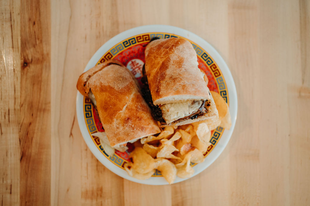 A ciabatta sandwich cut in half, filled with greens and a slice of cheese, is placed on a decorative plate. The plate also holds a serving of potato chips. The items are on a polished wooden table.