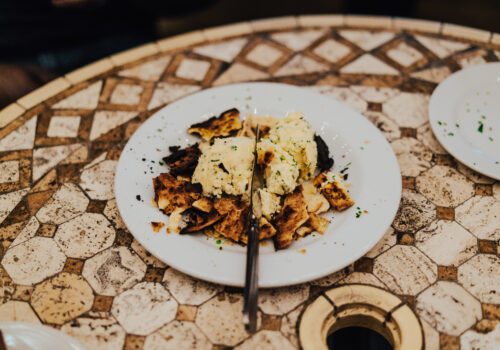 A plate of scrambled eggs garnished with chopped chives and served with pieces of crispy, toasted flatbread on a round, patterned table. There is a fork resting on the plate. Image