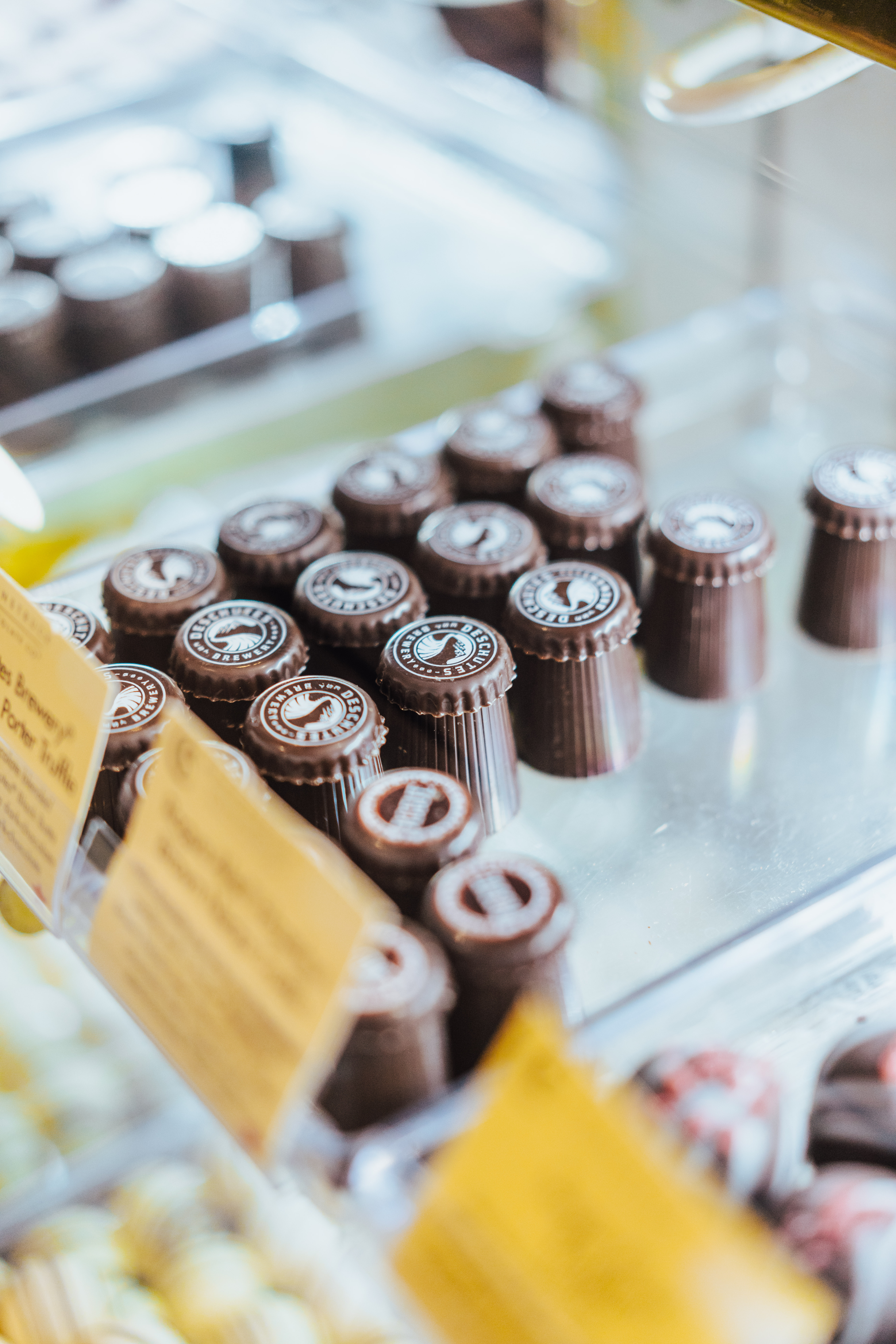 Close-up view of a display case filled with neatly aligned gourmet chocolates. The chocolates are circular with intricate designs on top. Labels with descriptions of the chocolates are partially visible in the foreground.