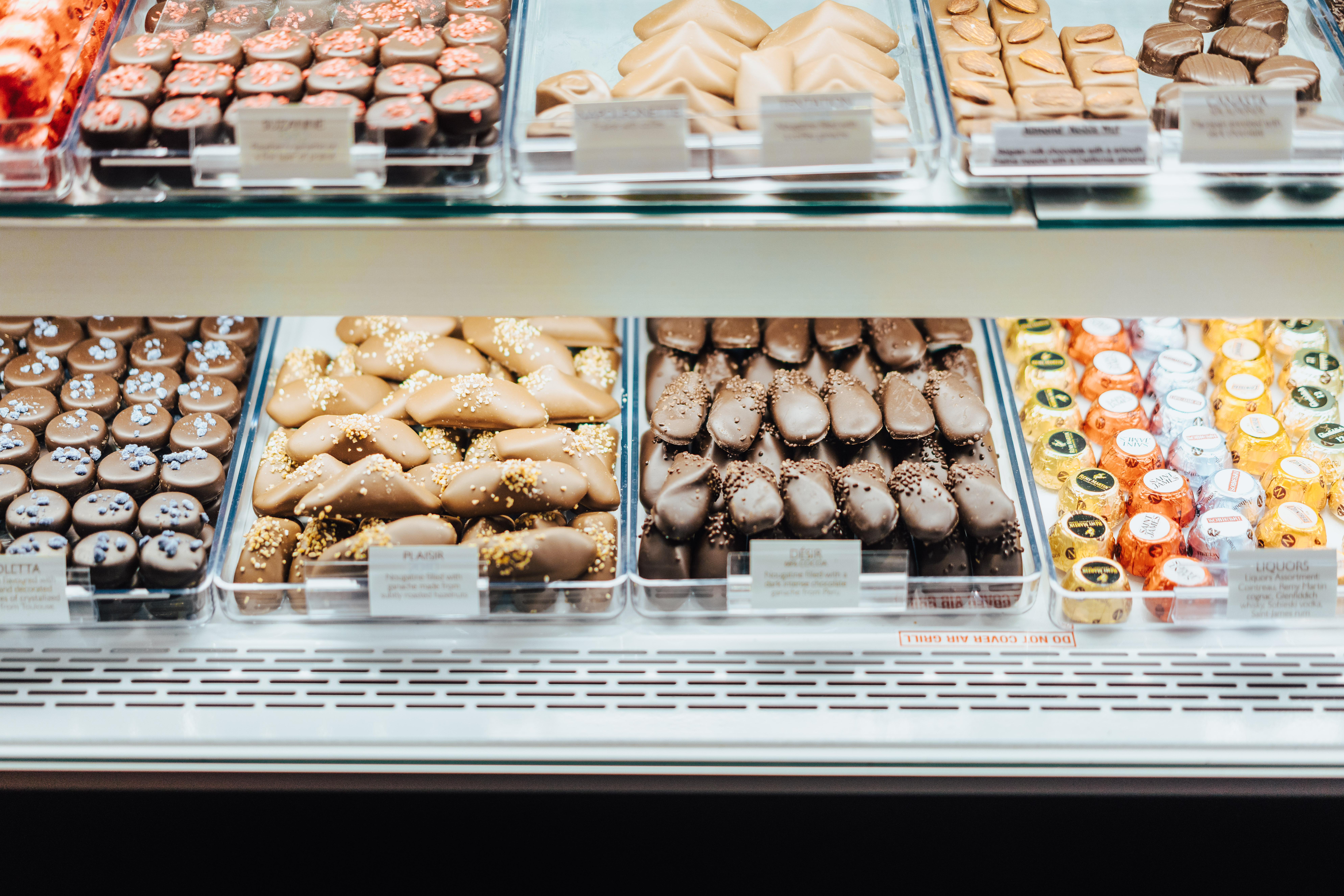 A display case filled with an assortment of chocolate confections, including dark chocolate pieces with sprinkles, milk chocolate squares, and chocolate-covered nuts. Small labels can be seen in front of each type of candy.